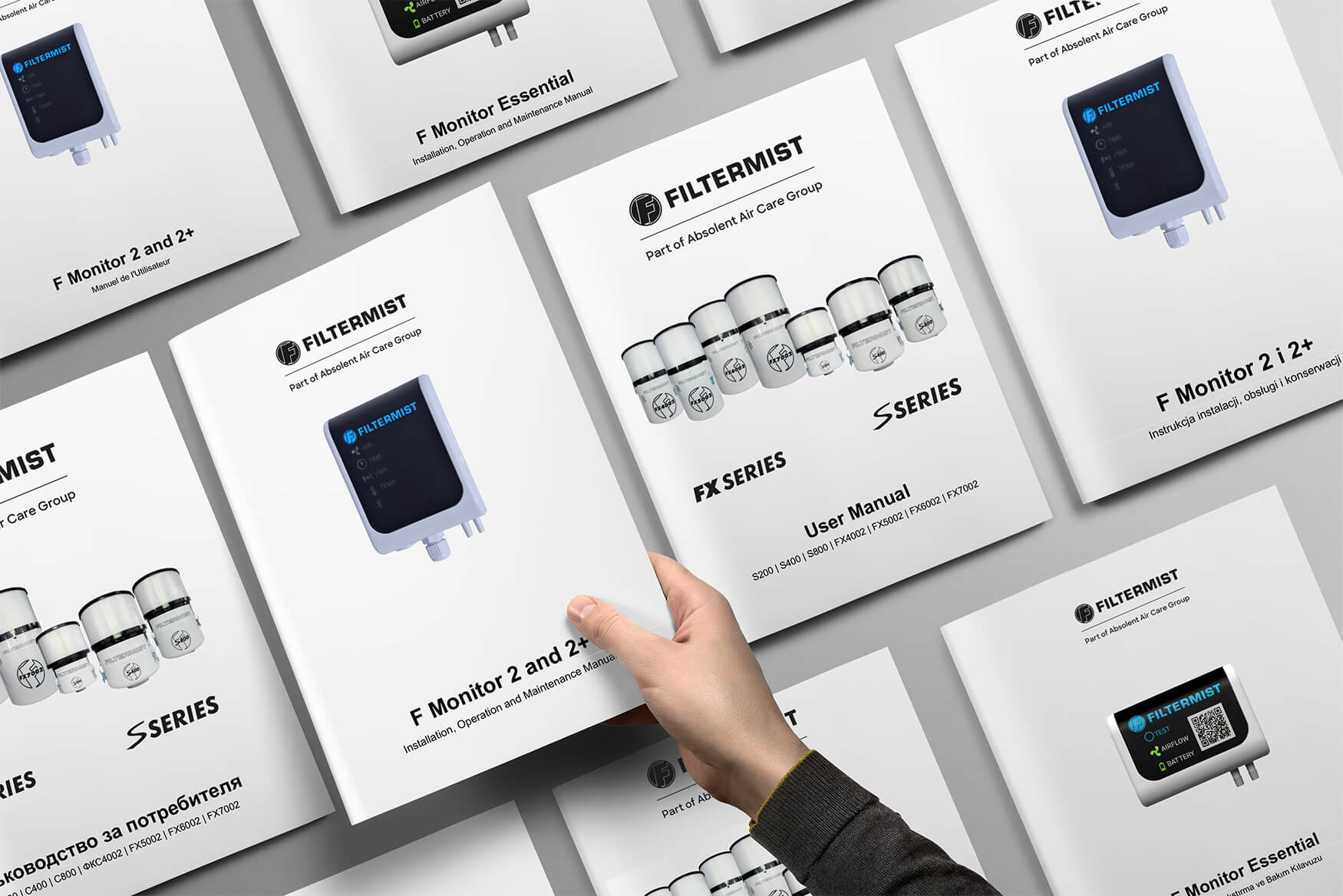 A hand reaching for a Filtermist user manual among several neatly arranged product manuals, featuring images of monitoring devices and filter products with clean, modern cover designs, all placed on a flat grey surface.